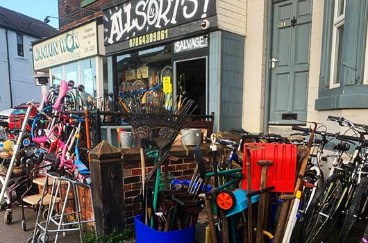 outside distance photo of allsorts secondhand thrift shop showing the lovely colourful logo in light green and amber coloured paintwork with a large glass display window and many items outside including bikes scooters gardening tools stools and toys