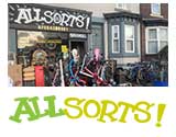 logo of all sorts  shop outside photo showing glass window and large luminous green and yellow logo and various items like bikes and scooters outside